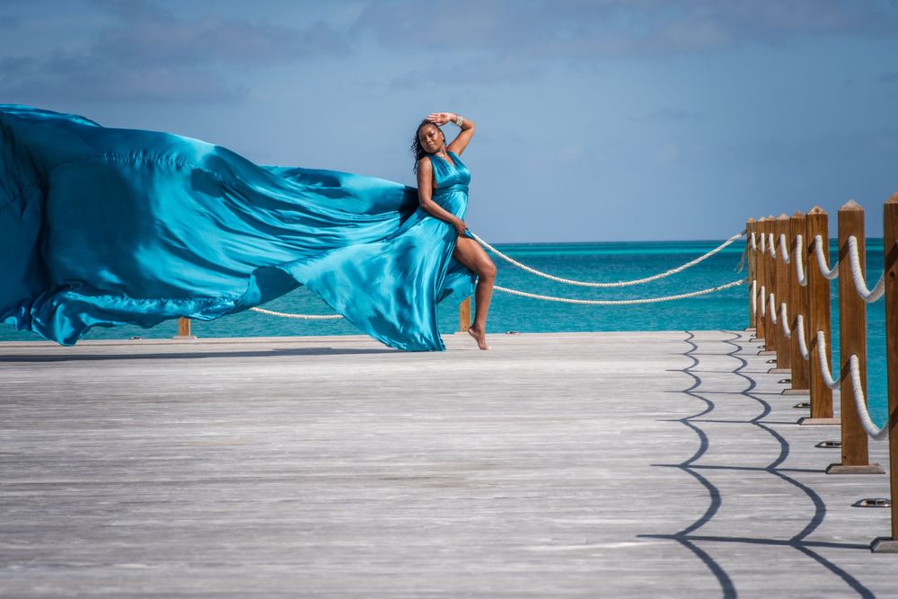 Woman in flowing teal dress jumping on pier with fabric billowing in tropical ocean breeze.