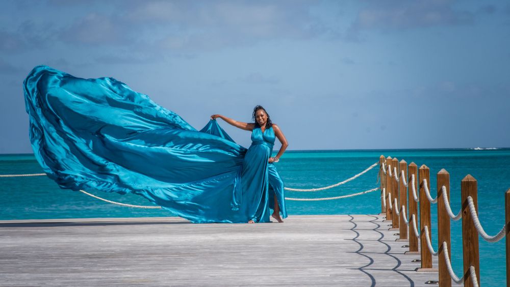 Elegant woman posing in teal flying dress on seaside pier with turquoise water backdrop.