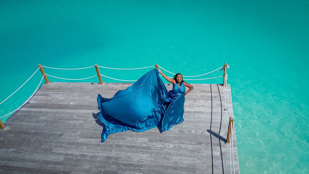 Aerial view of woman in blue satin dress on wooden pier surrounded by turquoise ocean water.