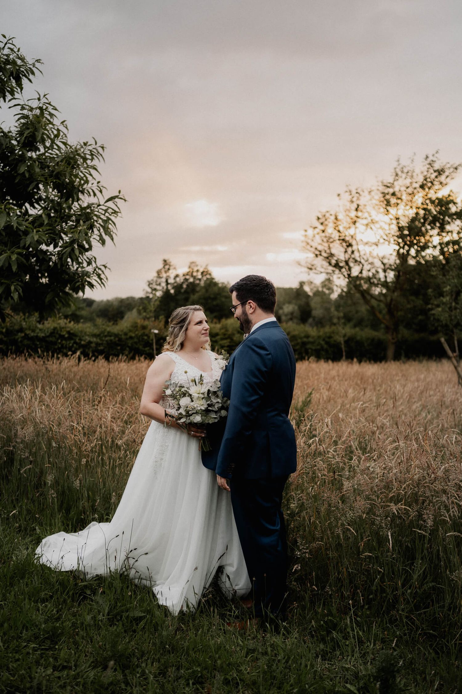 A couple stands in a golden field at sunset for their wedding photos, with the bride in white and groom in navy blue.
