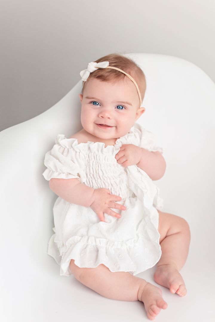 Baby in white smocked dress and bow headband on white chair.