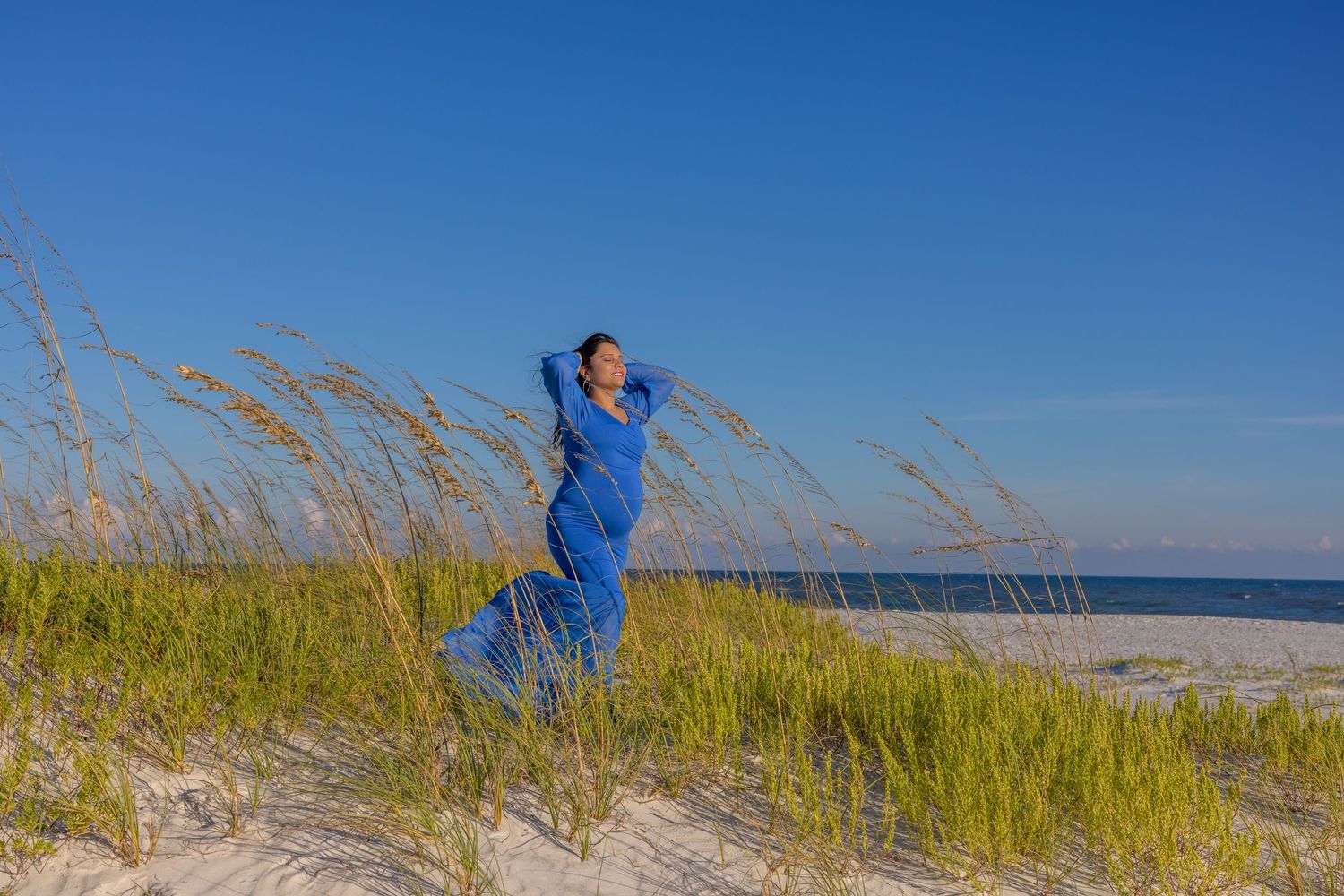 Person in blue dress standing among beach grass against bright blue sky.