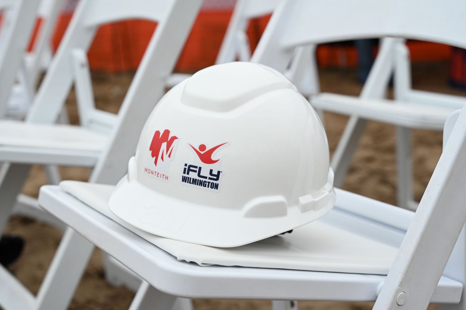 White construction hard hat with red logo sitting on folding chair.