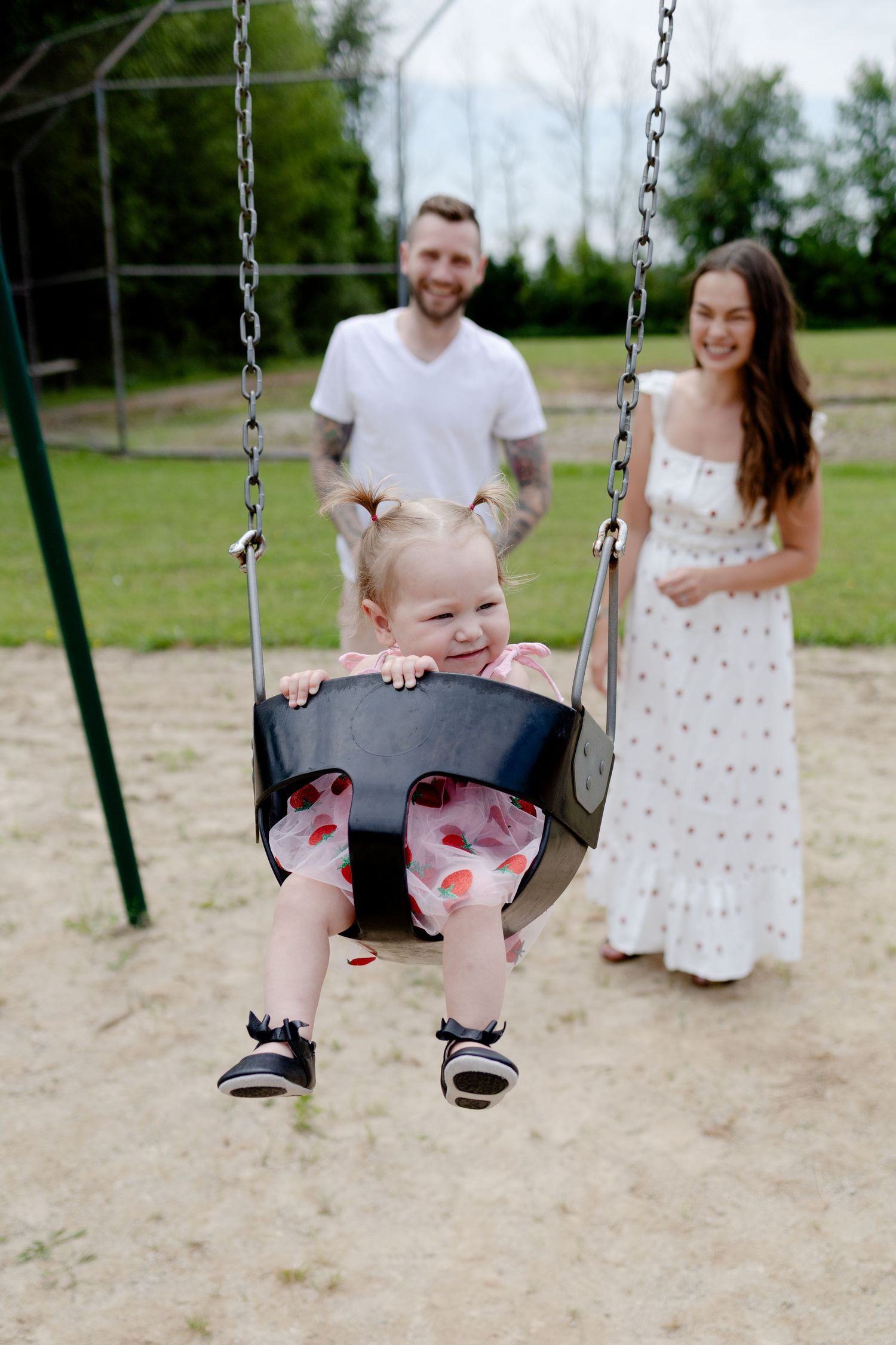 a husband and wife laughing and pushing their daughter on the swing during a family photoshoot in the park