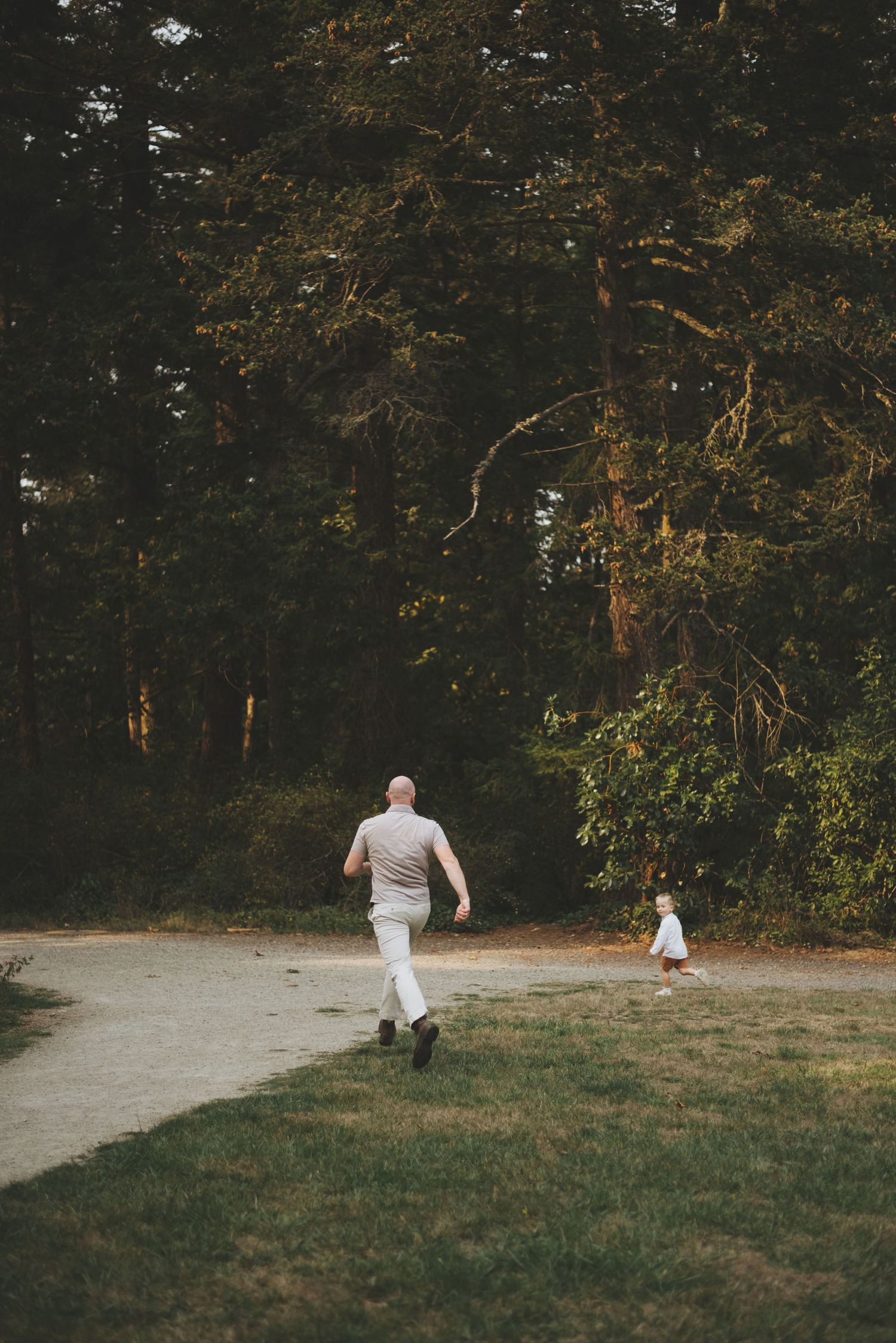 Two figures running along a wooded path surrounded by tall evergreen trees.