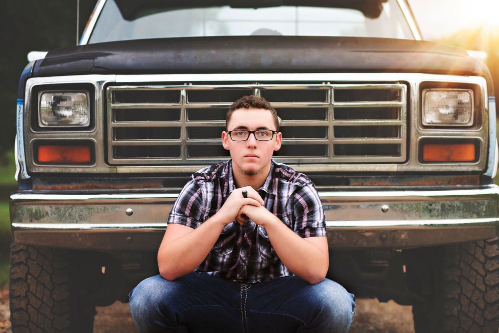 Man in plaid shirt sitting on vintage truck with sunset glow.