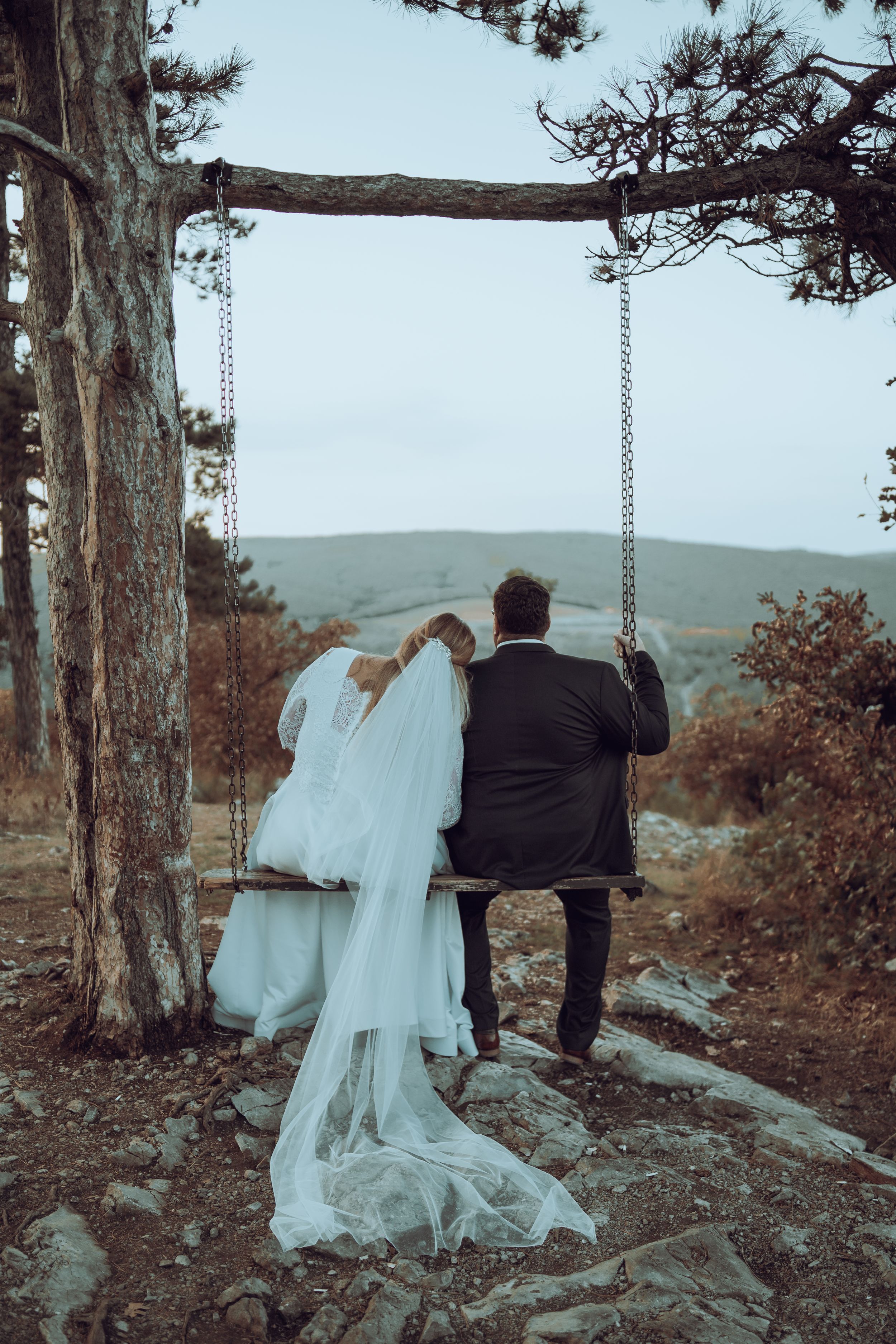 A couple on a rustic wooden tree swing enjoy a scenic mountain view, with a flowing white wedding dress trailing behind them.