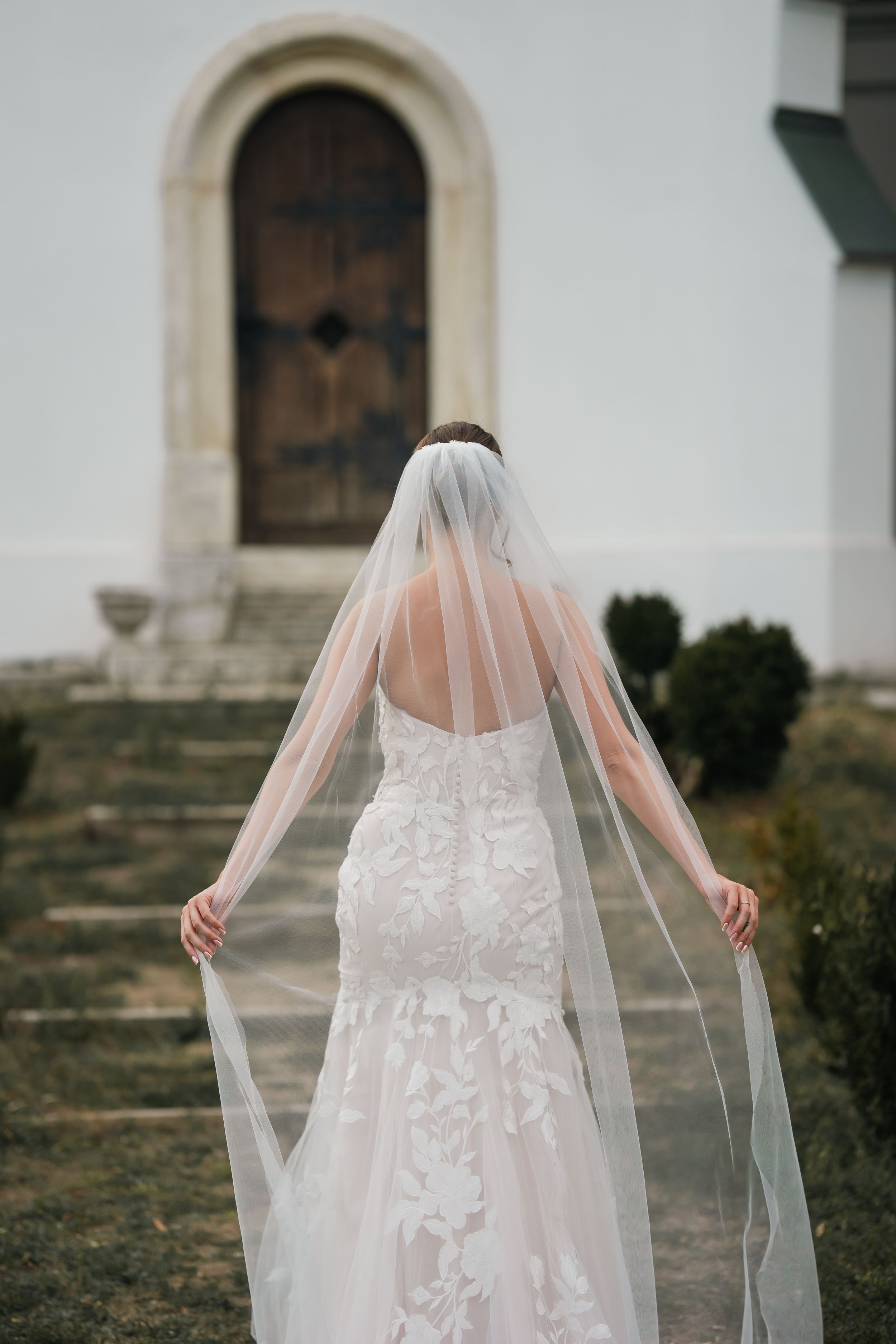 A bride in a lace wedding dress stands in front of a church door while holding her flowing veil out to both sides.