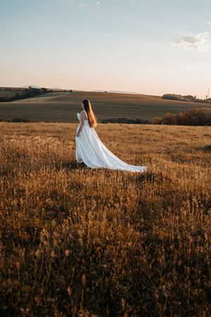 Egy lebegő fehér menyasszonyi ruha elegánsan lengedez egy aranyló búzaföldön naplementekor, álomszerű vidéki környezetben.