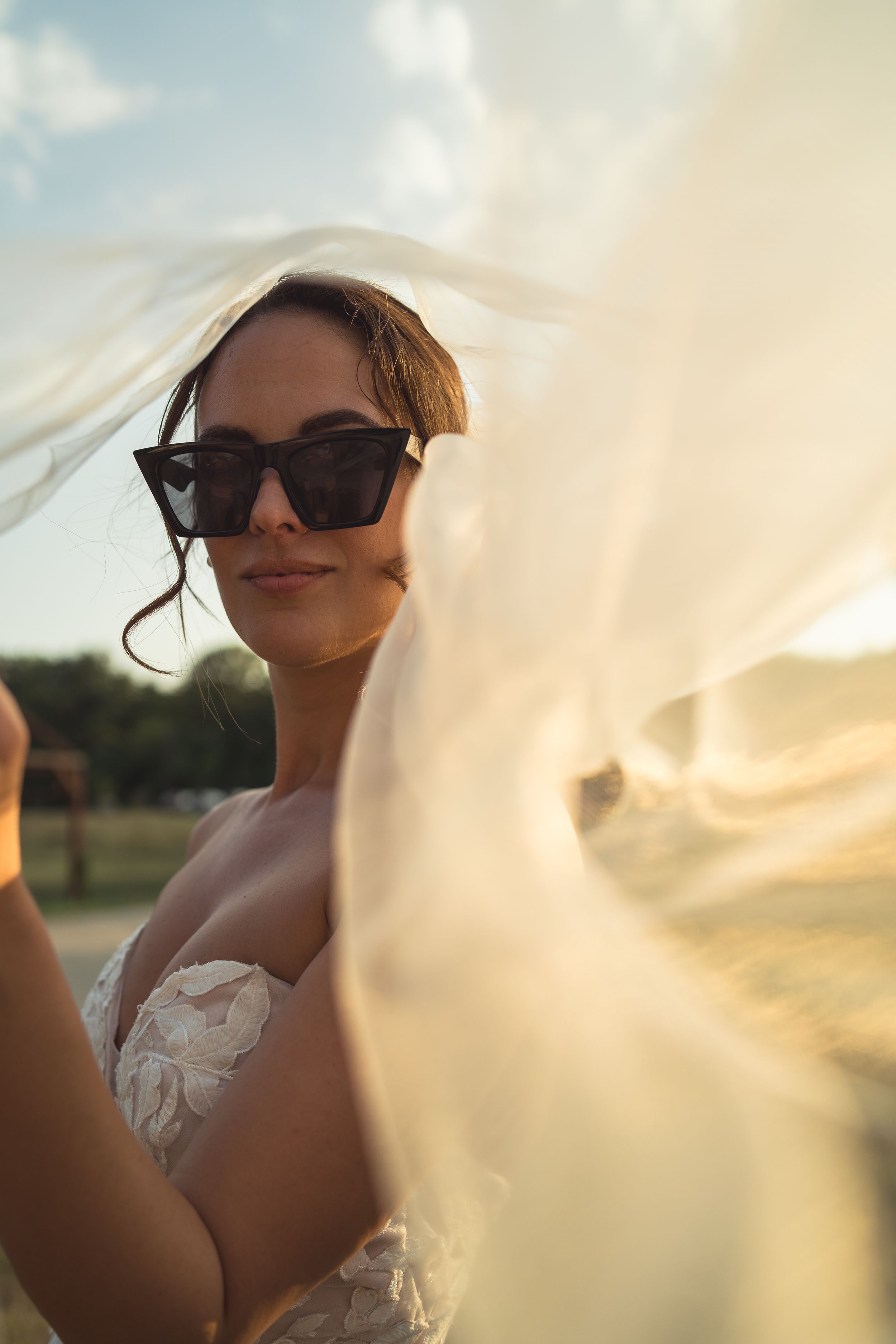 A bride wearing sunglasses and a strapless wedding dress poses outdoors at sunset with a flowing veil.