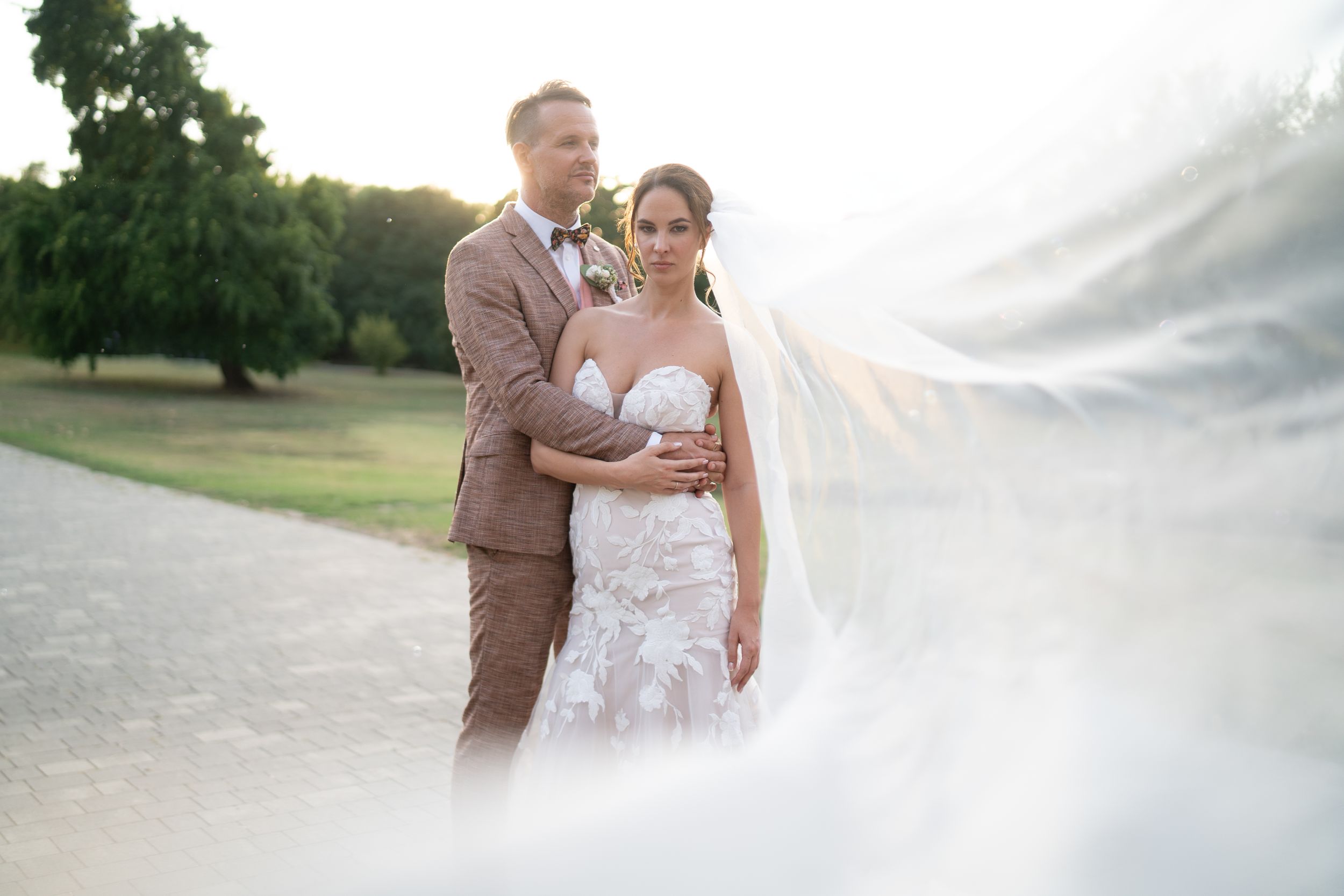 A beautiful wedding couple poses outdoors on a path with a flowing veil catching the sunlight at golden hour.