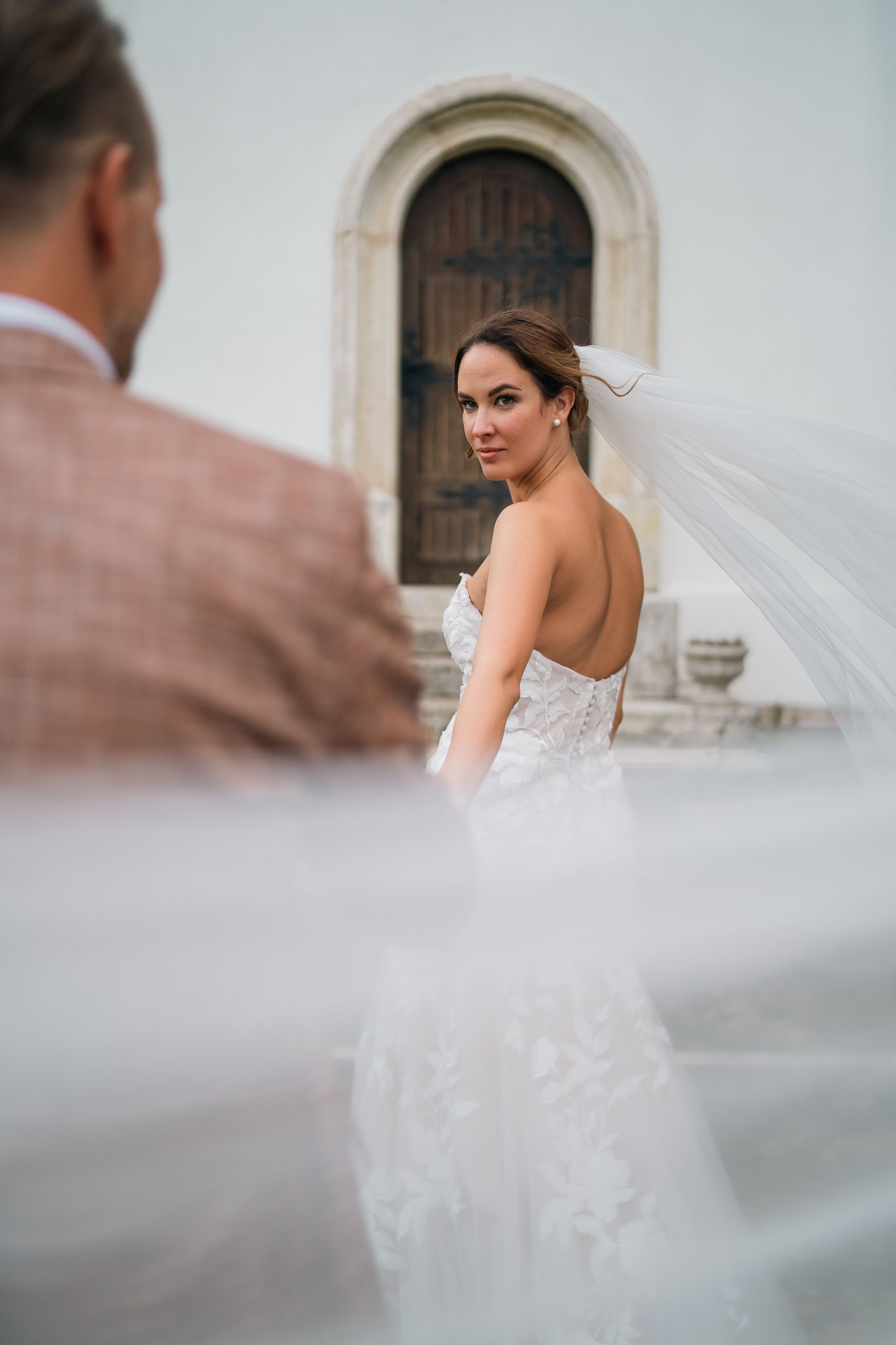 A bride in a strapless lace wedding gown with flowing veil stands in front of an arched wooden door at a church.