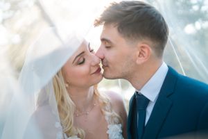 Egy romantikus pár gyengéd esküvői csókot oszt meg egy lebegő fehér fátyol alatt, lágy természetes fényben.