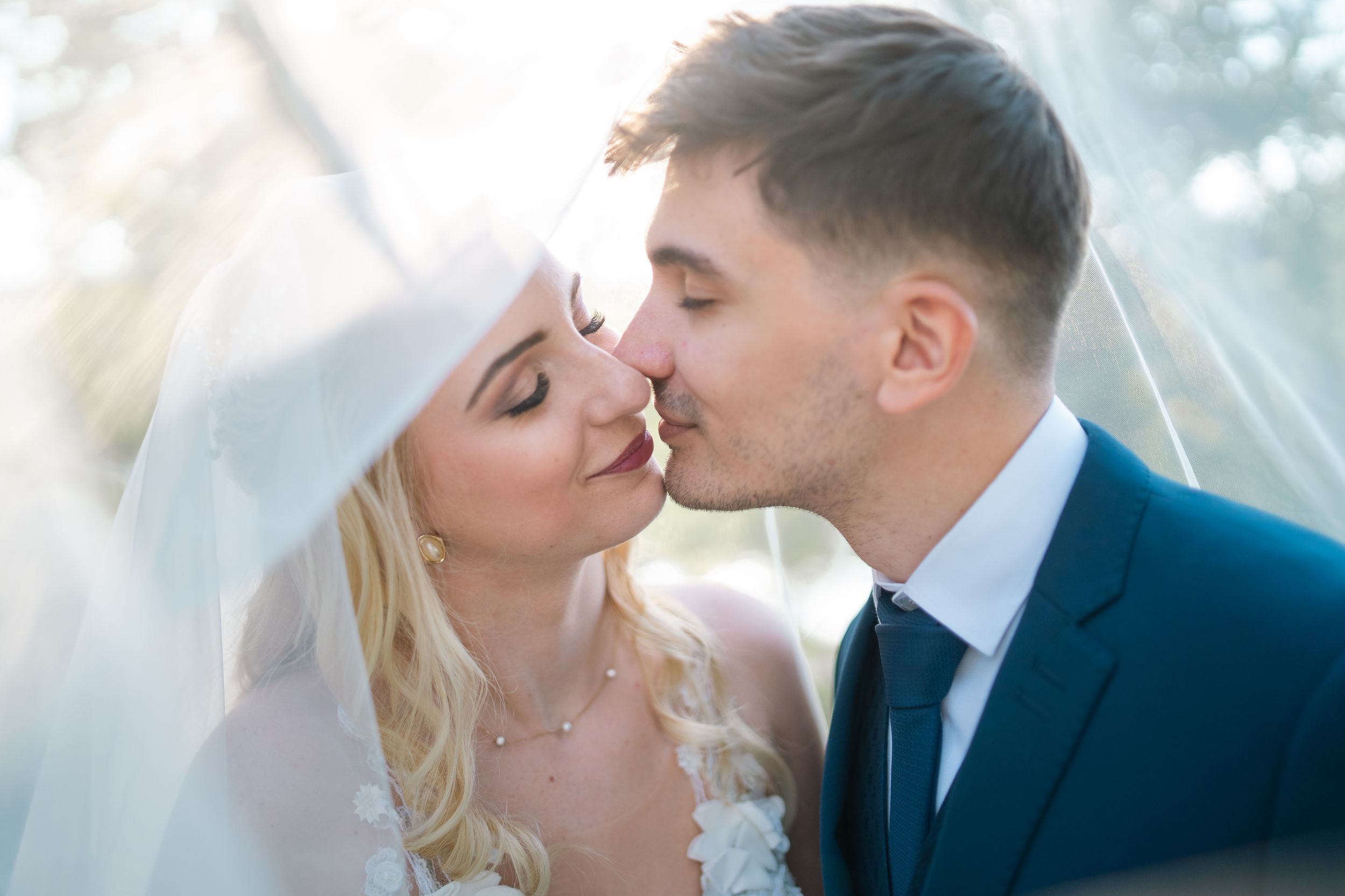 A romantic couple sharing a tender wedding kiss under a flowing white veil in soft natural lighting.