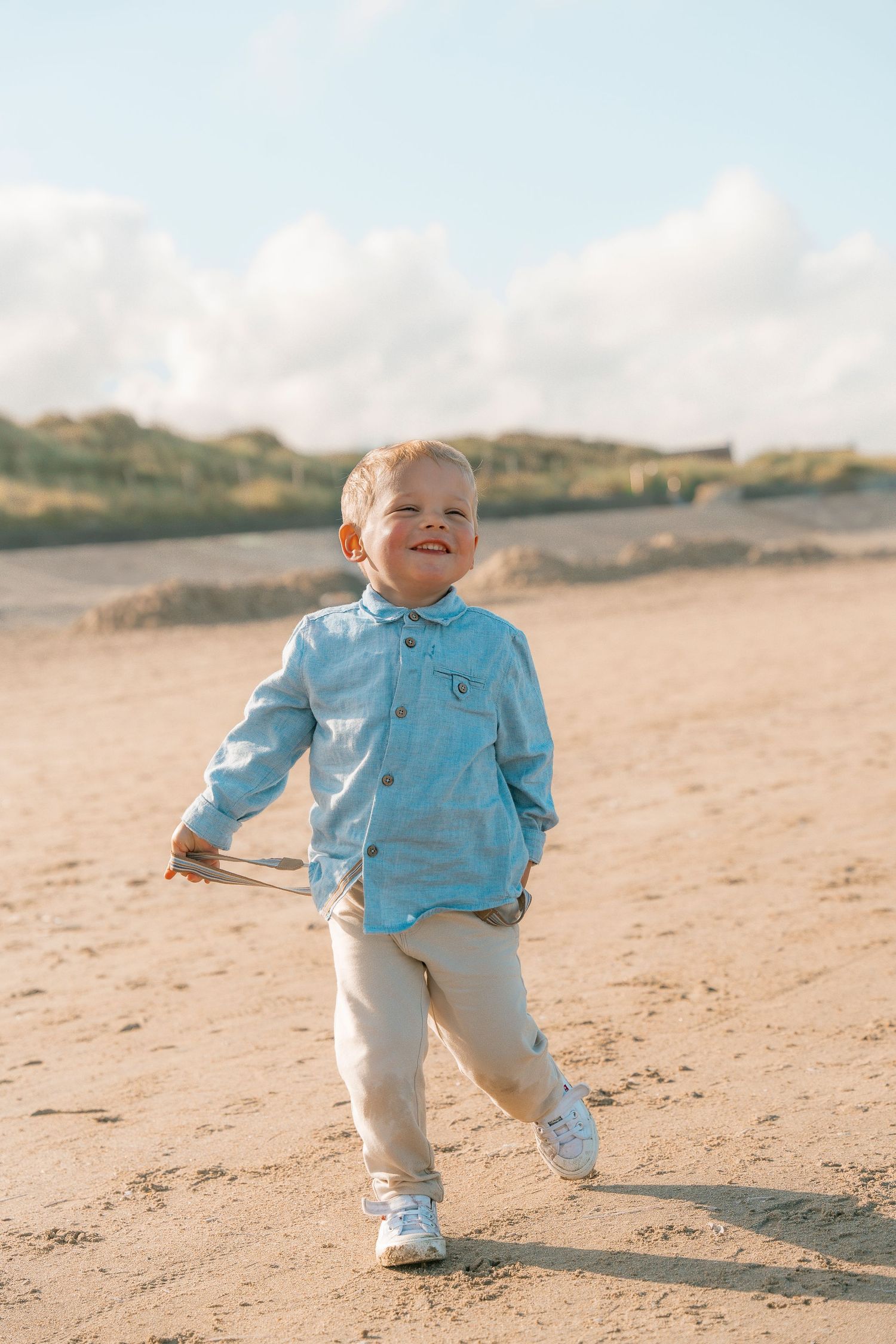 Young child in blue jacket and white pants running joyfully on sandy beach.