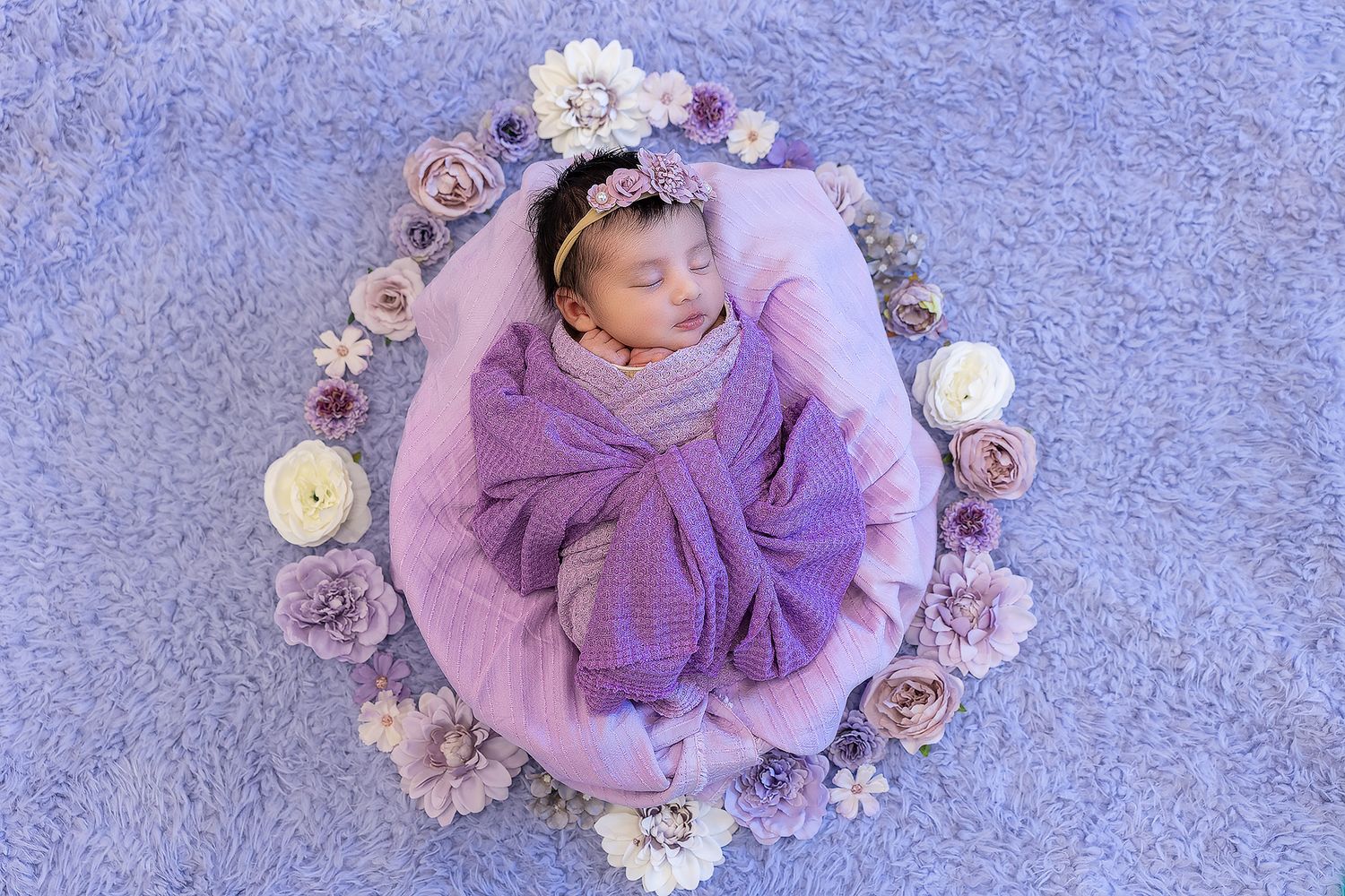 Sleeping newborn wrapped in purple swaddle surrounded by pink and purple flowers on blue backdrop.