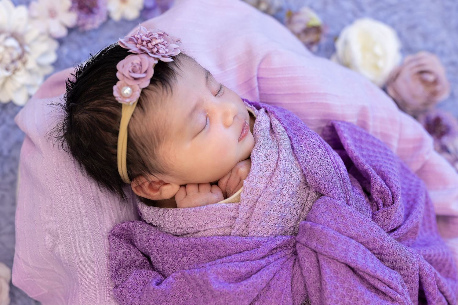 Newborn baby sleeping peacefully in purple wrap with floral headband.