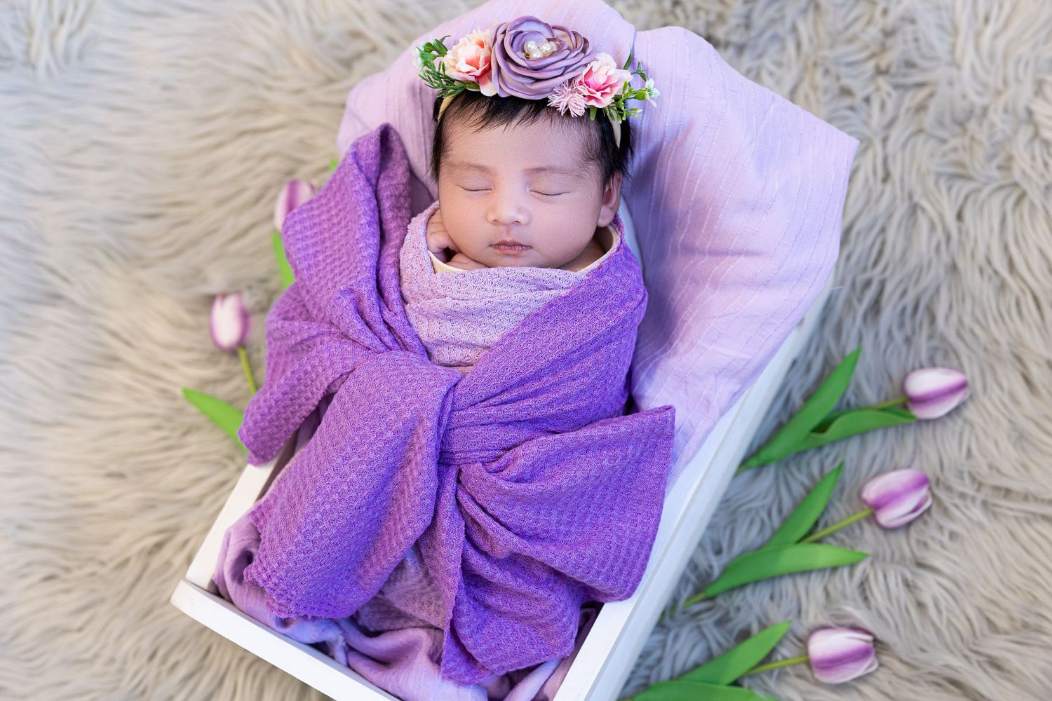 Newborn baby sleeping in white basket with purple wrap and floral crown surrounded by tulips.