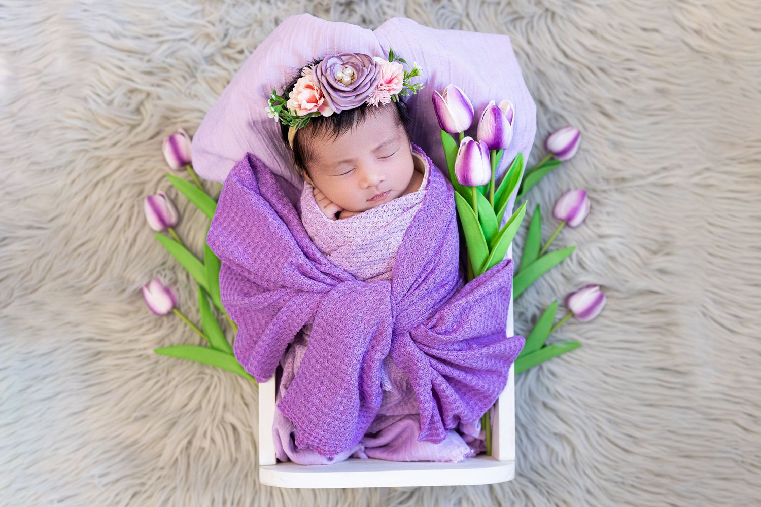 Sleeping baby in purple wrap among pink tulips and flowers on furry background.