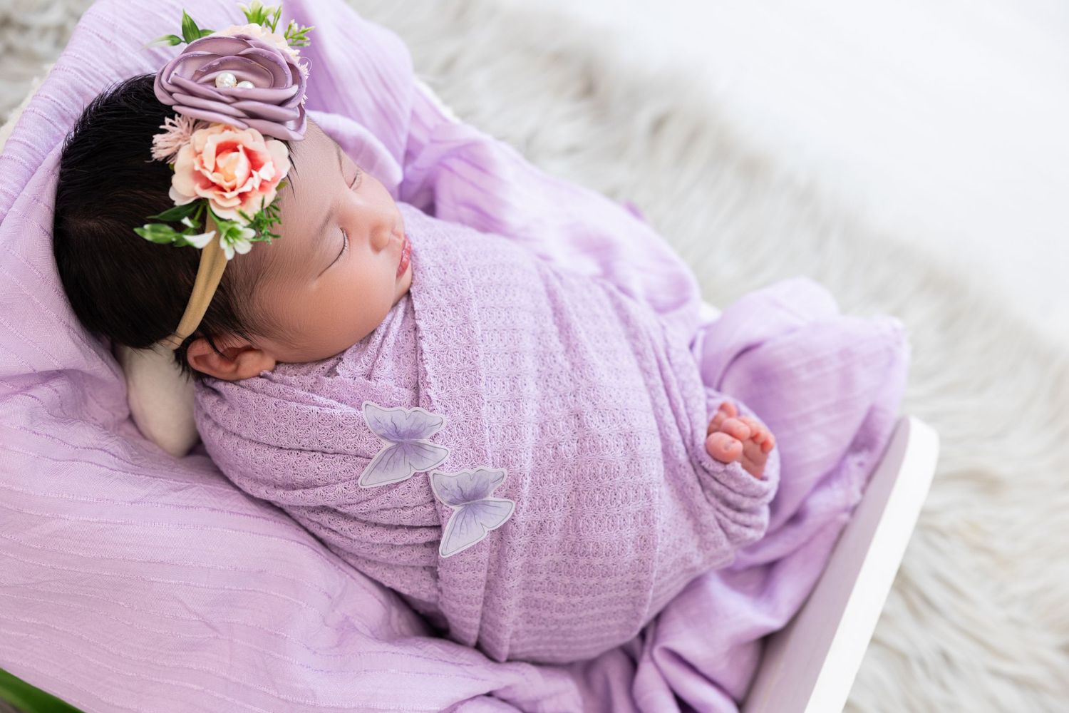 Peaceful newborn sleeping in purple wrap with floral crown on soft pink background.