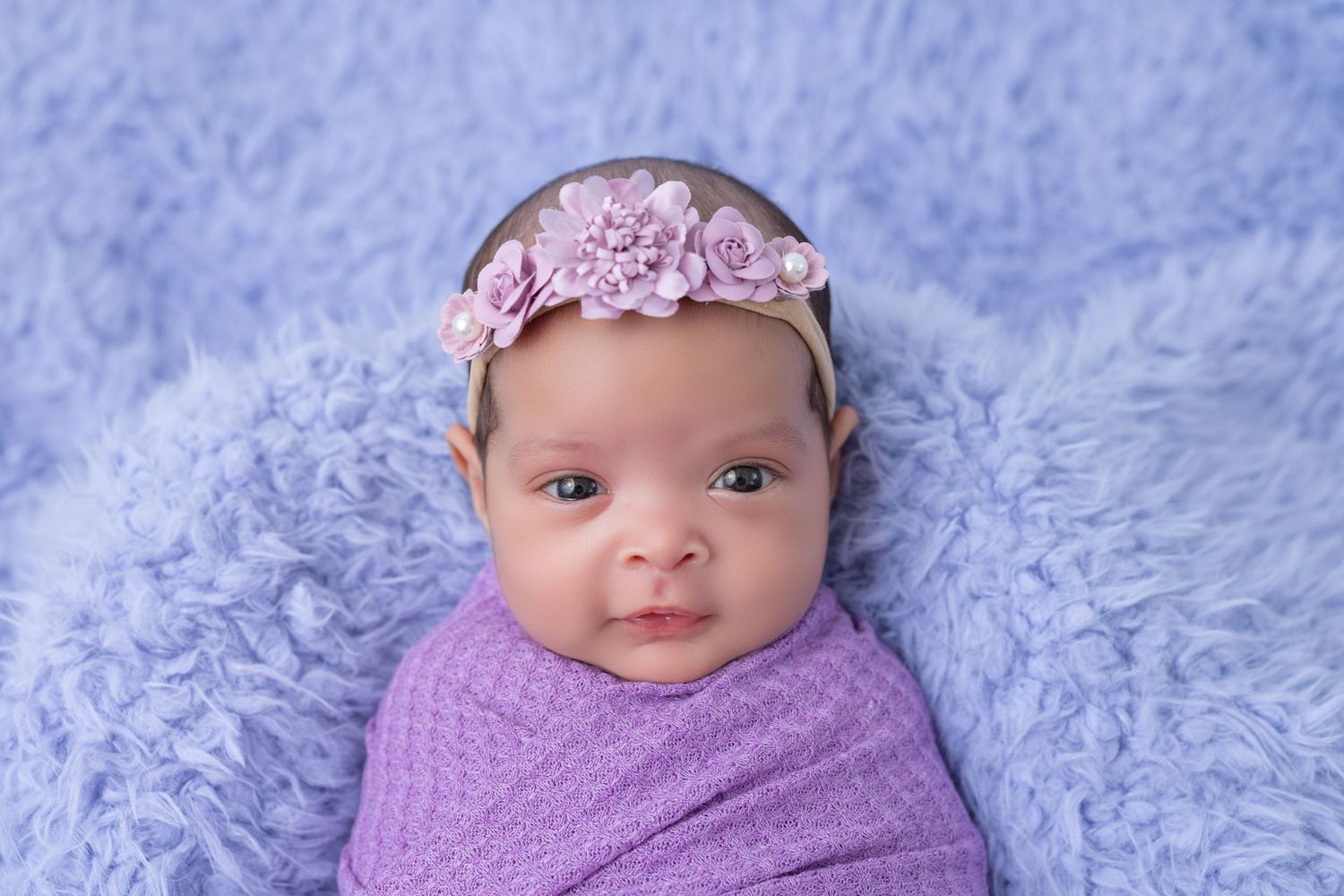 Baby wearing purple headband wrapped in purple blanket on textured blue background.