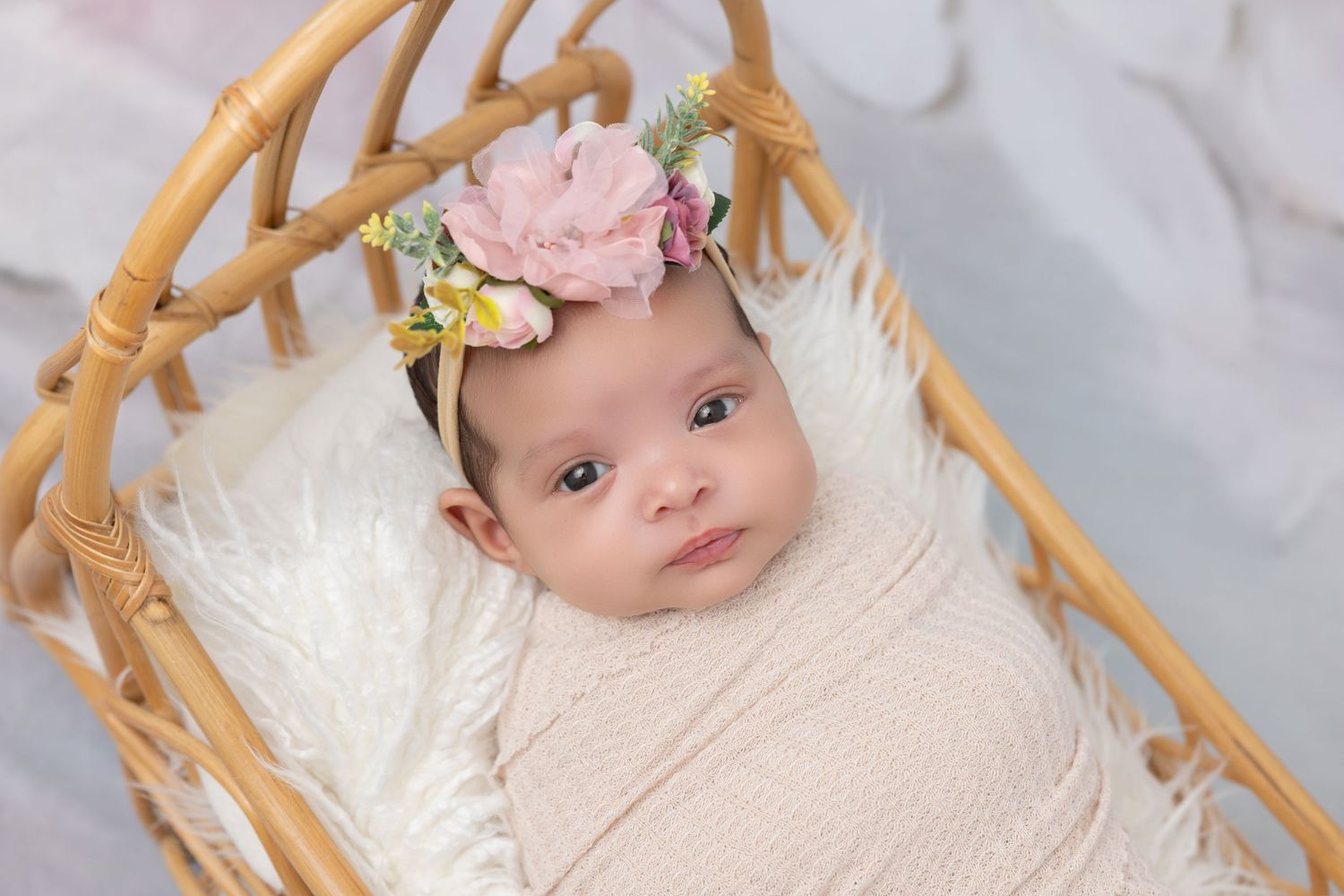 Newborn in woven basket wearing floral headpiece and cream wrap.