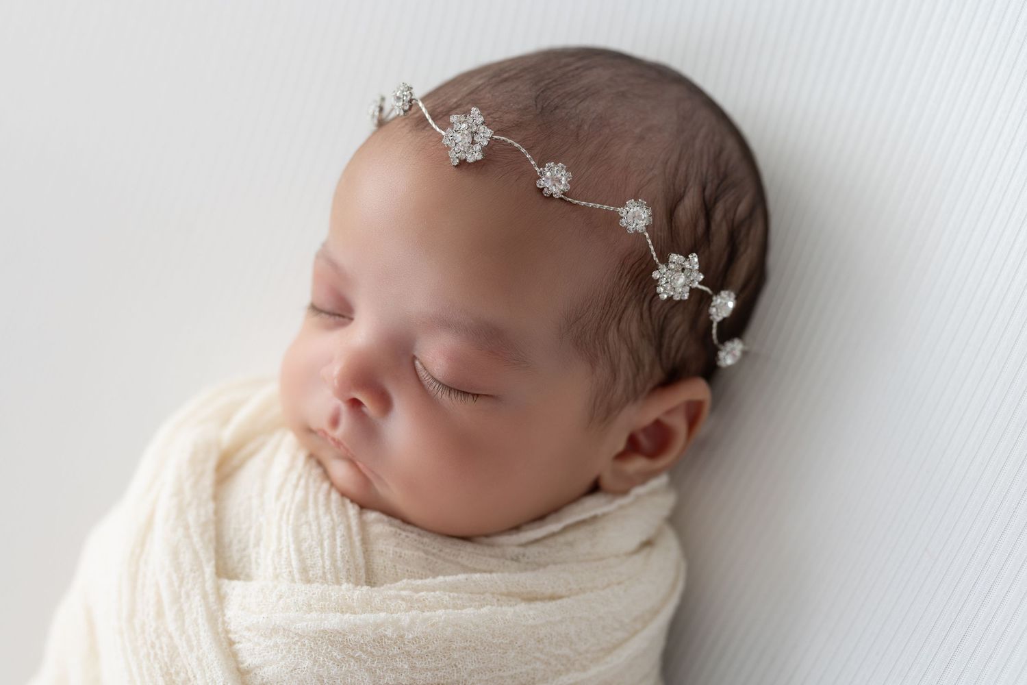 Sleeping newborn wearing delicate crystal headband wrapped in cream blanket.