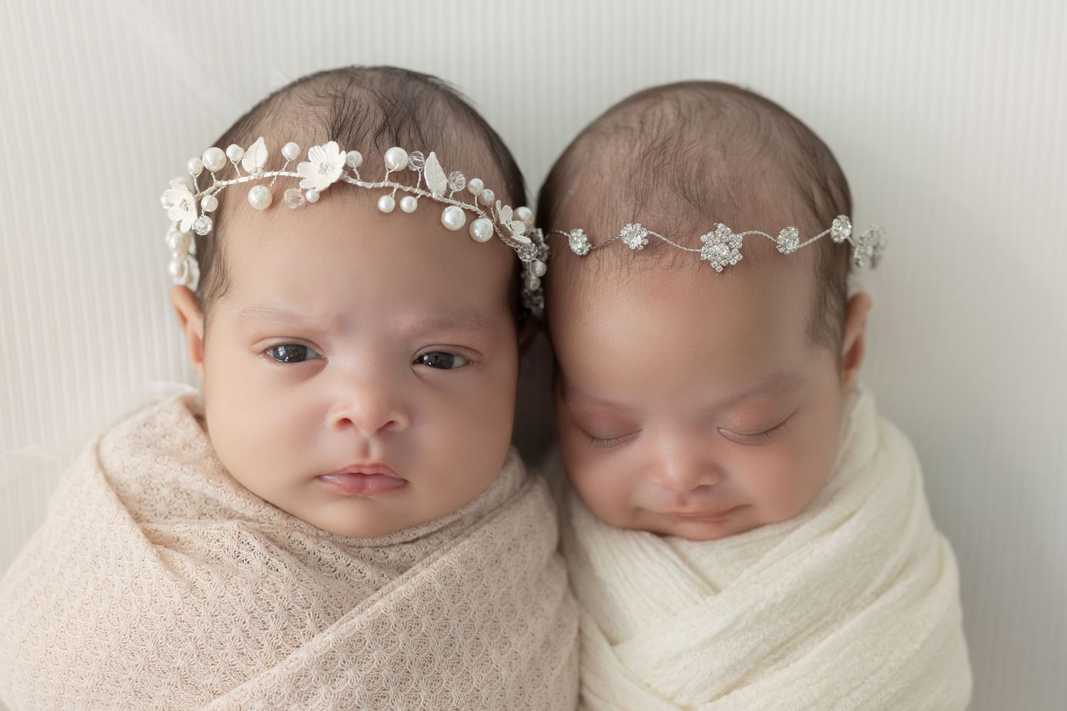 Twin newborns swaddled together wearing matching crystal headbands.