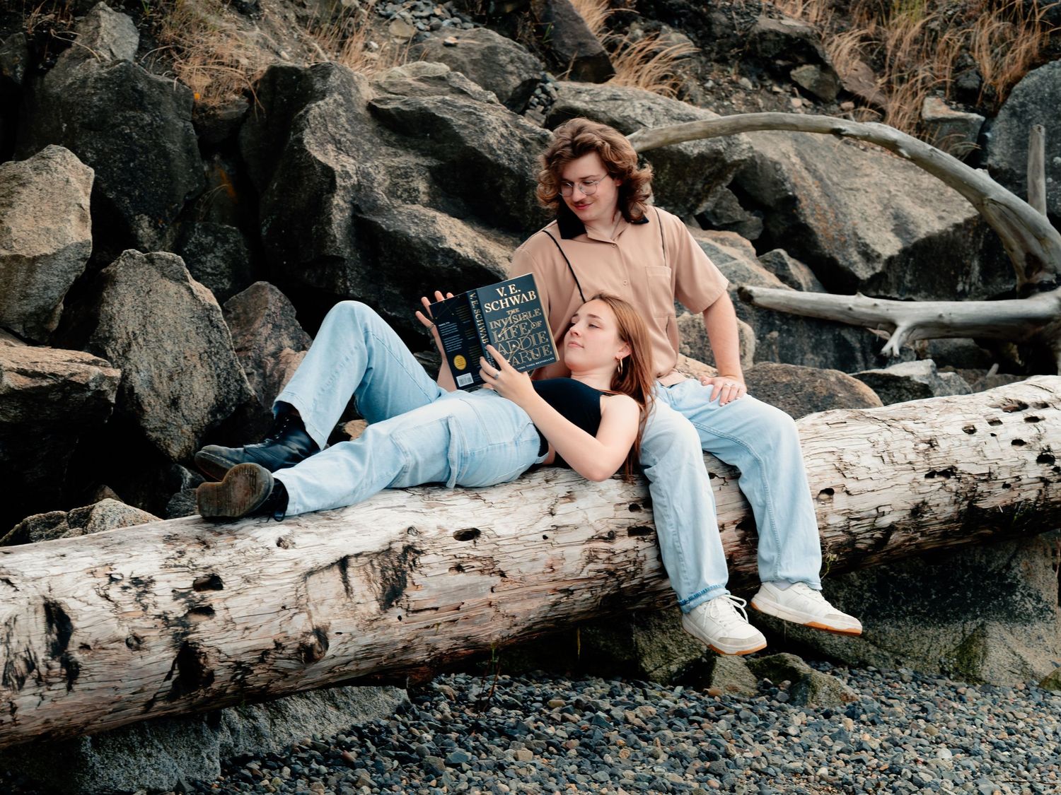 A couple lying on a large piece of driftwood at a rocky beach in casual jeans and white sneakers.