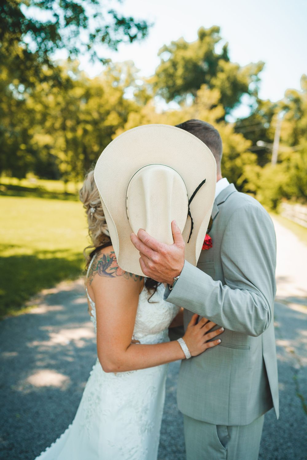 Playful wedding moment with couple kissing behind a cowboy hat on a sunny path.