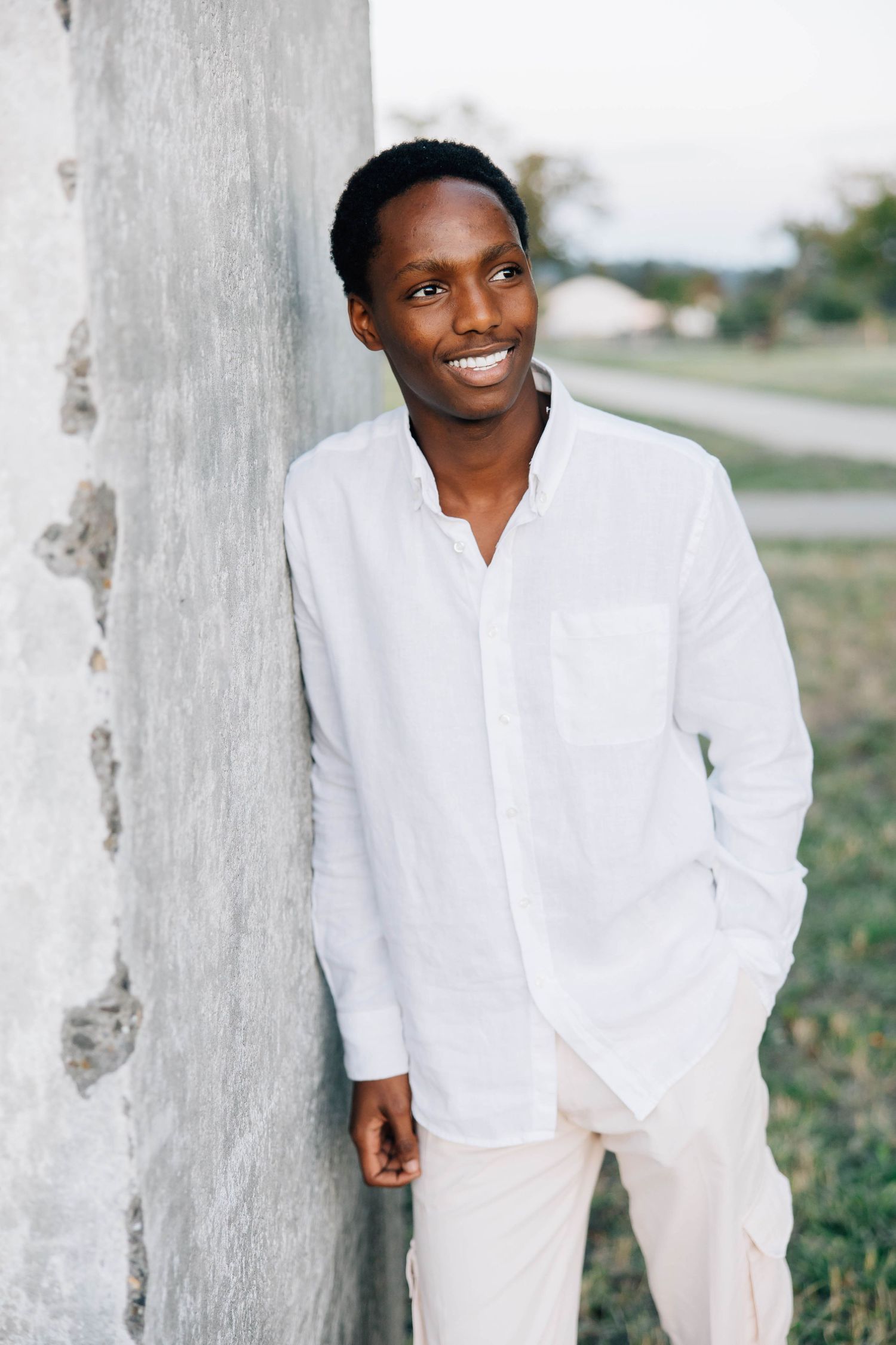 Confident young Black man in white linen shirt leaning against concrete pillar outdoors.