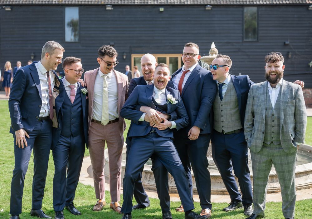Groomsmen in stylish suits pose together for a fun group photo outside a rustic barn.