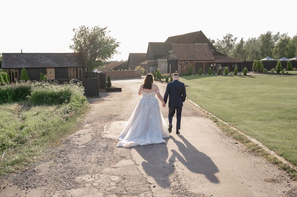 Newlyweds walking hand in hand down a path towards a rustic wedding venue.