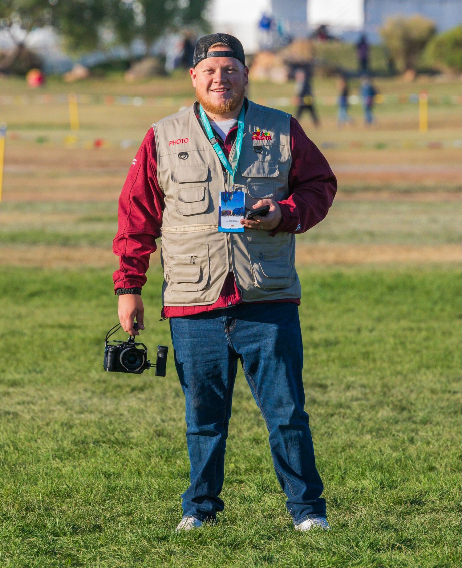A photographer wearing a utility vest holds camera equipment while standing in a grassy field.