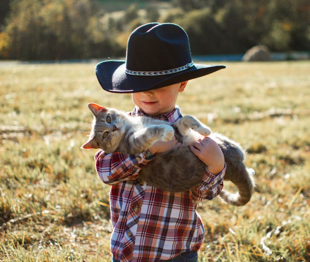 child wearing cowboy hat holding a cat in a grass field
