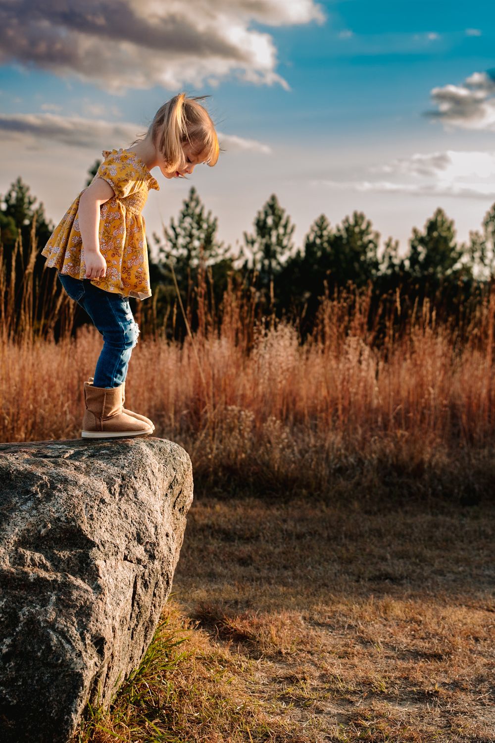 Young child stands on large boulder against sunset sky and autumn landscape.