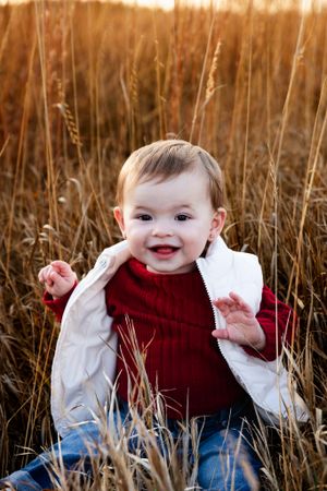 Small child in red sweater and white vest sitting among tall grass at golden hour.