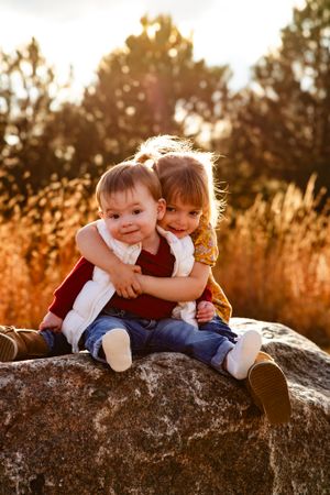 Two children share a warm embrace while sitting on a large rock during sunset.
