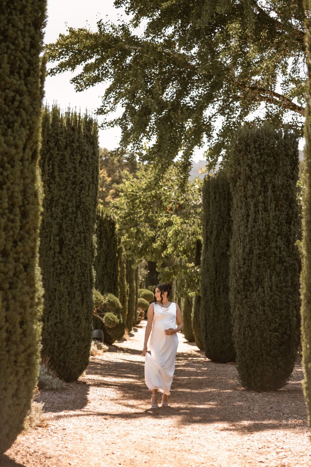Bride in flowing white dress walks through elegant cypress tree-lined path.