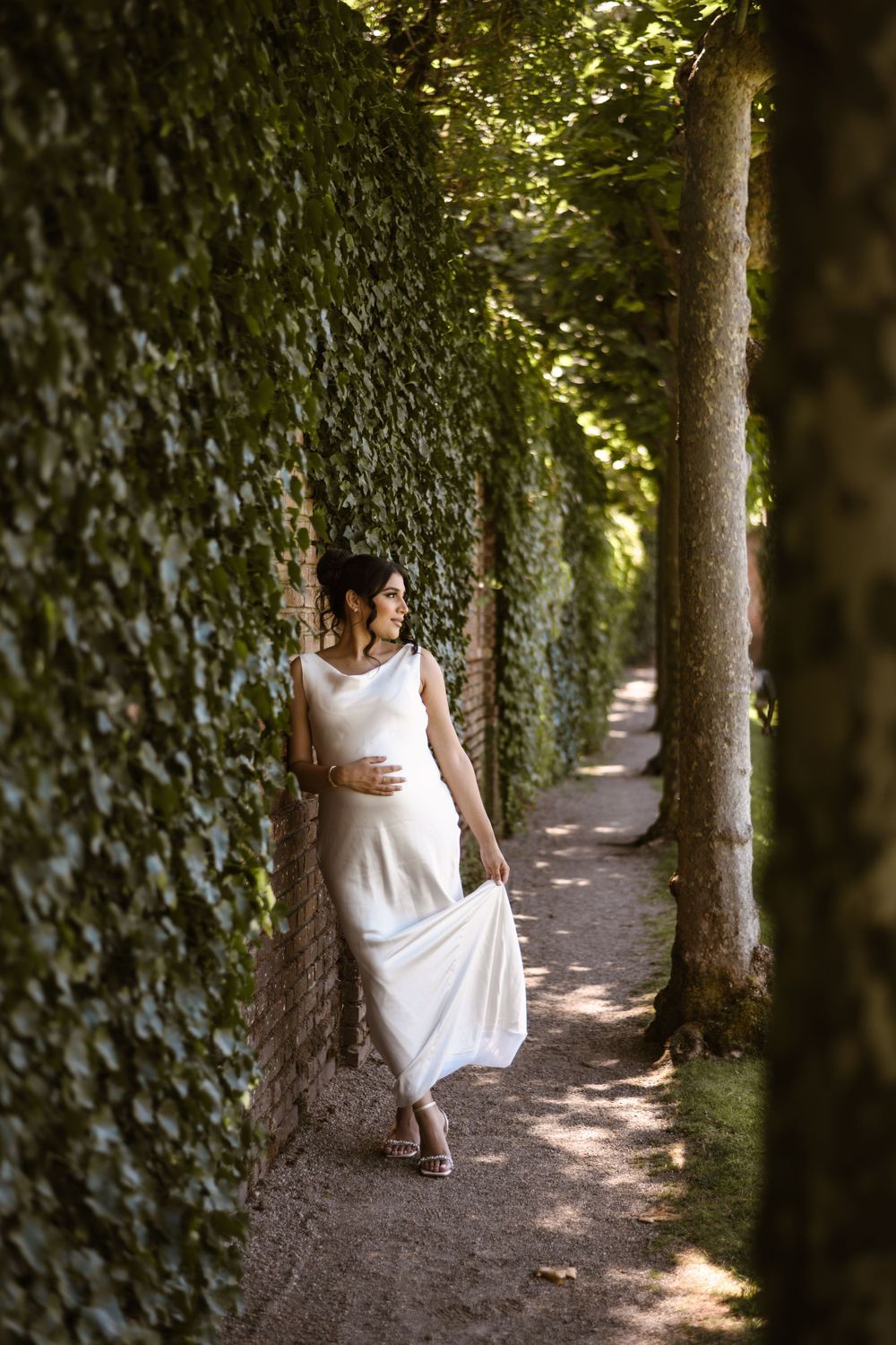 A white dress flows elegantly through a tree-lined garden path dappled with natural sunlight.