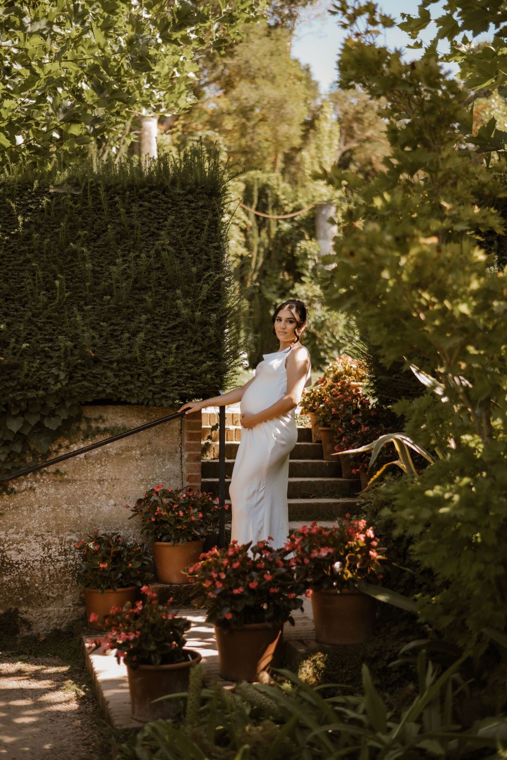 A white dress flows elegantly through a sunlit garden path with brick steps and potted flowers.