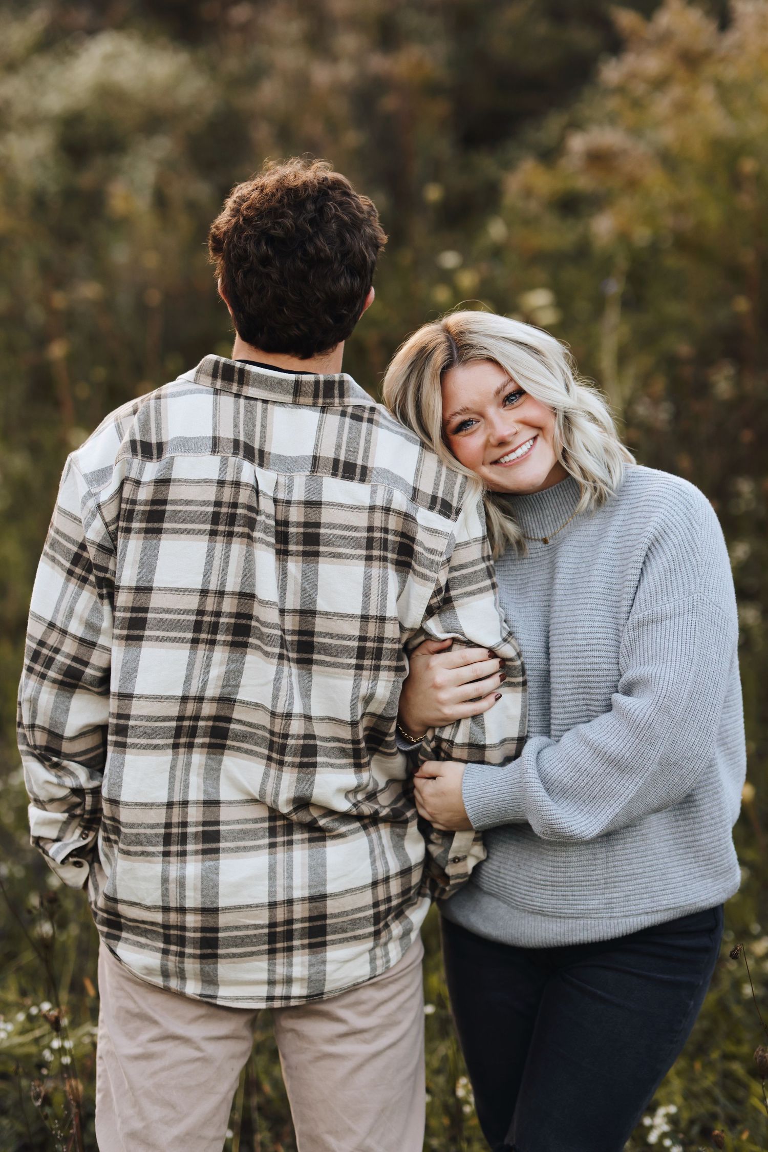 A couple embraces outdoors, with one wearing a plaid flannel and the other in a cozy gray sweater in an autumn setting.