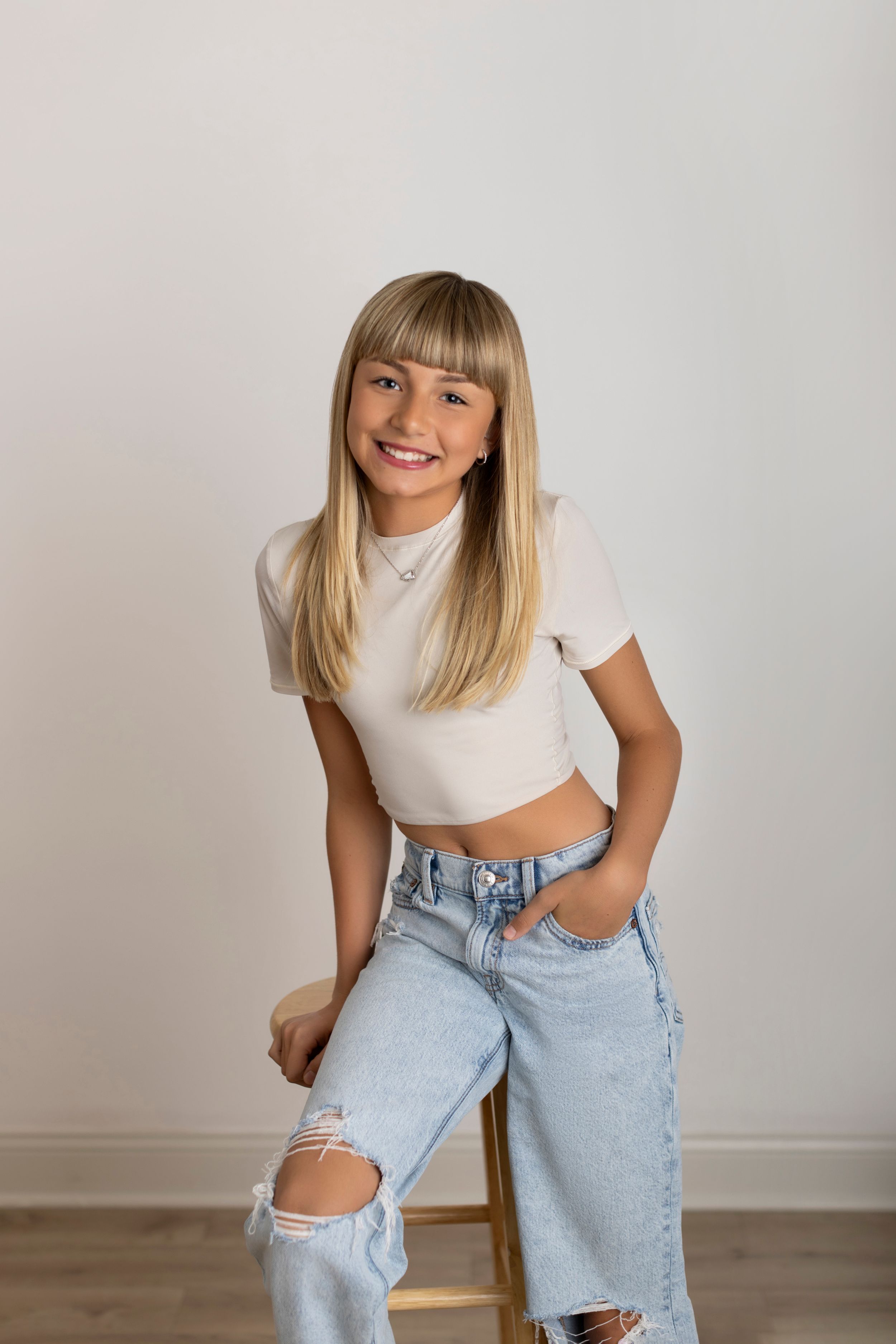 Person in white crop top and light wash ripped jeans poses against white wall in casual fashion shoot.