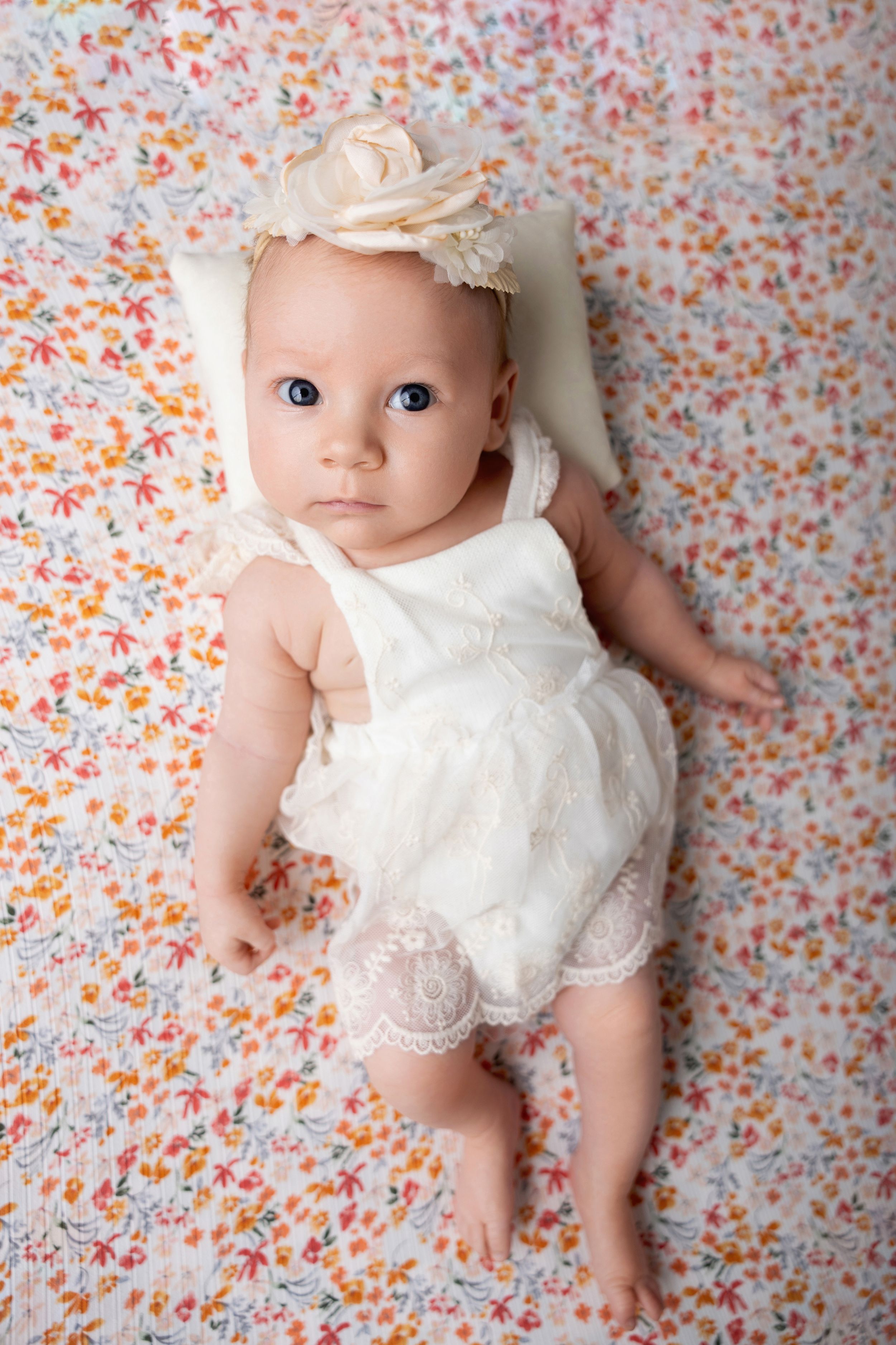 Baby lying on floral background wearing white lace dress and headband in series of newborn photos.