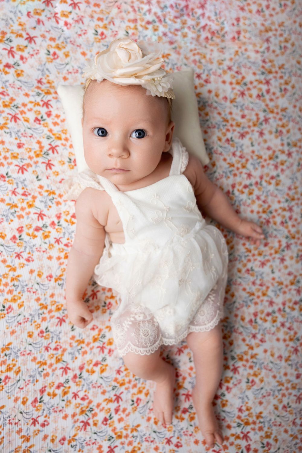 Baby lying on floral background wearing white lace dress and headband in series of newborn photos.