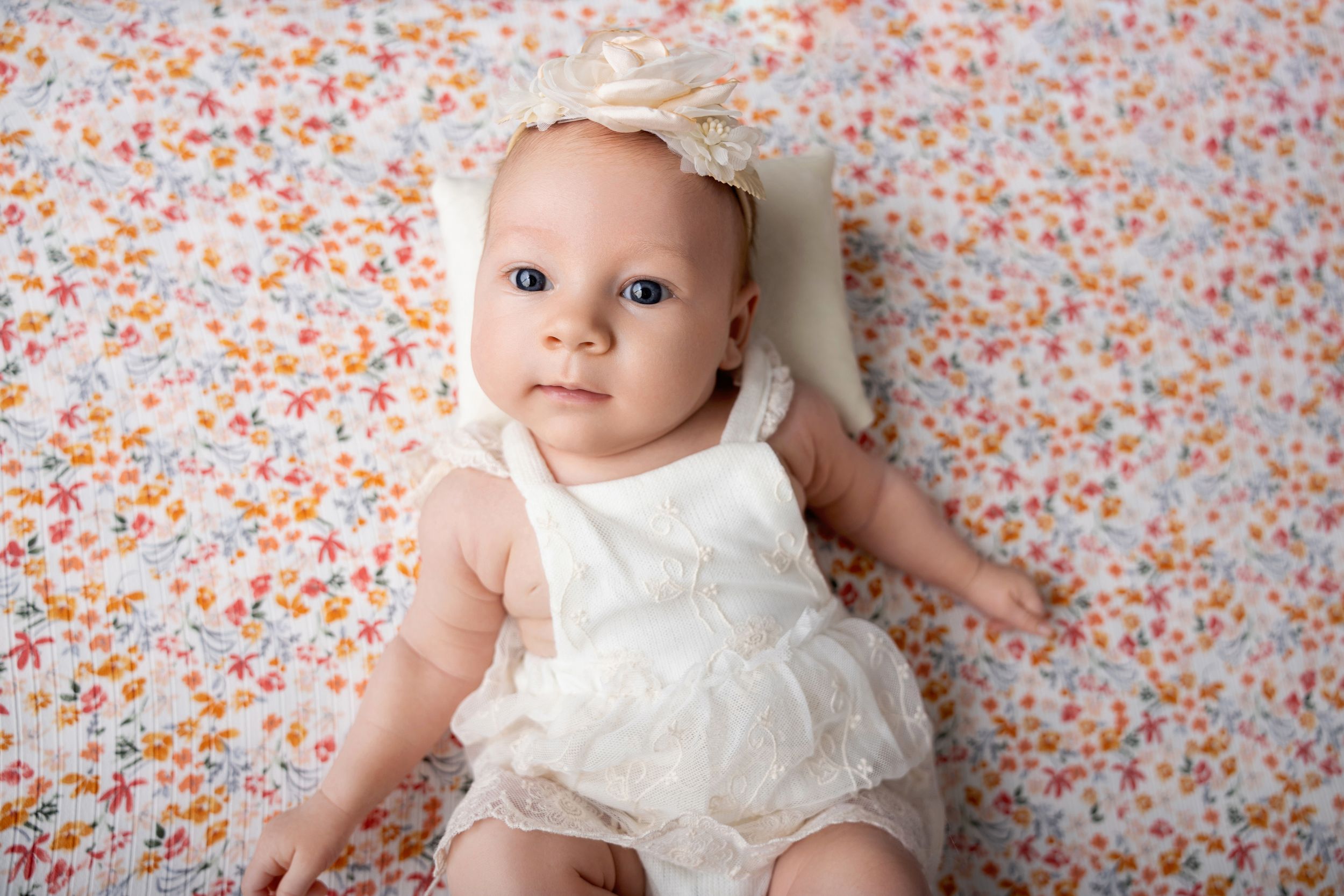 Small baby in white lace romper lying on floral patterned background with soft peachy colors.