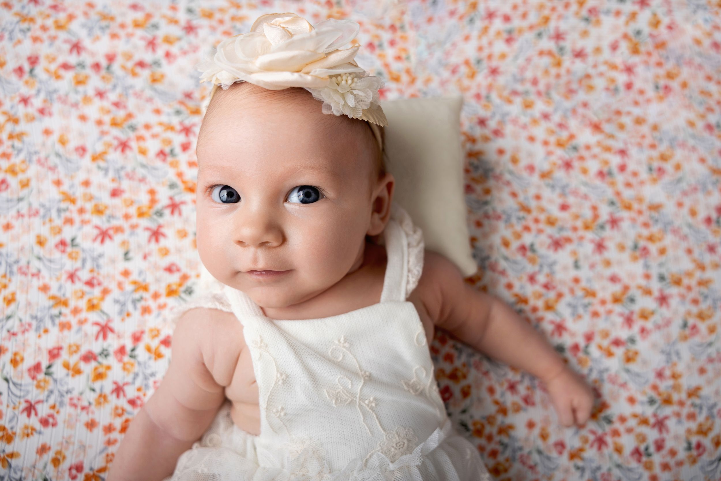 Baby in white dress lying against floral background smiling at camera.