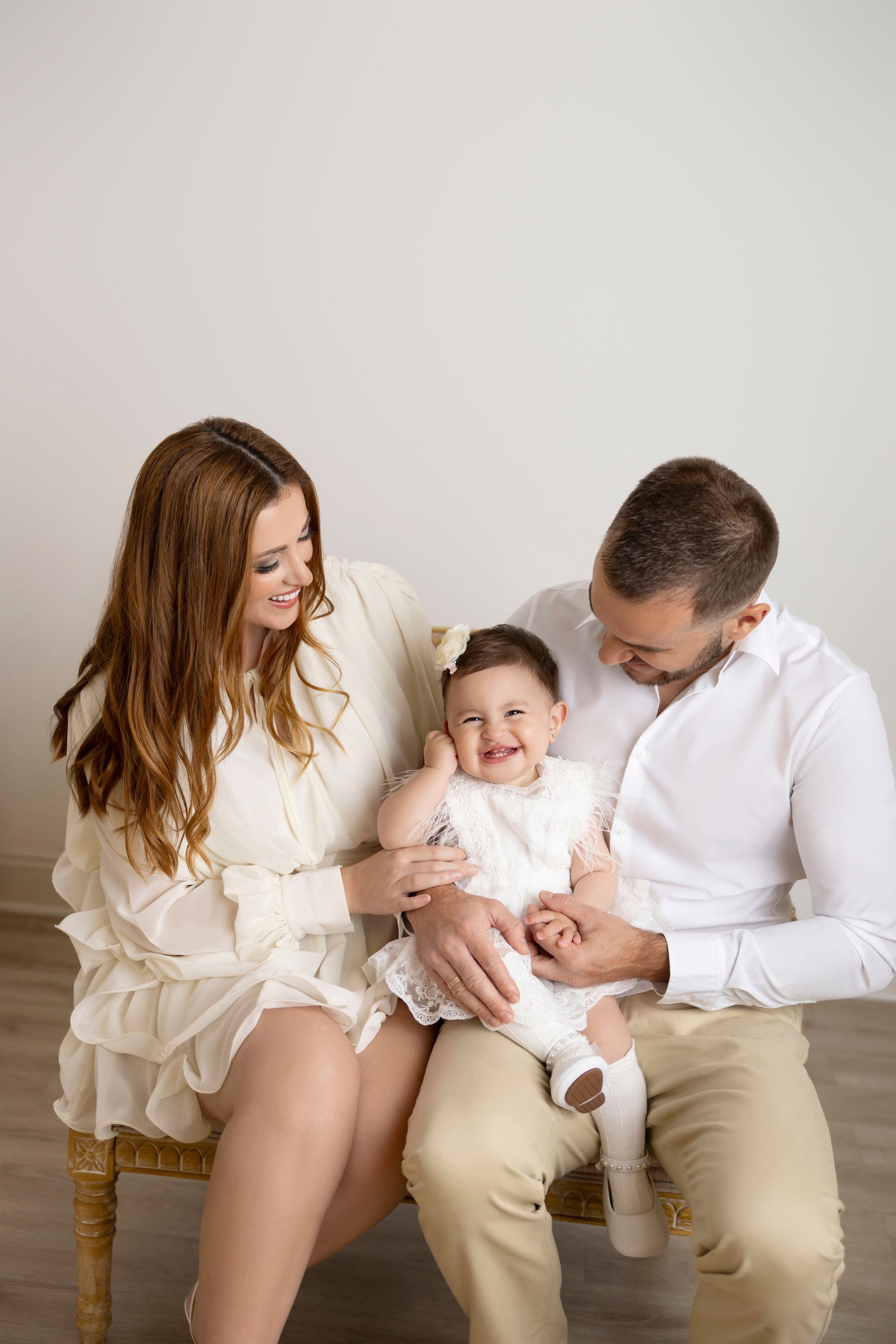 A family wearing white and beige outfits shares tender moments together in a minimalist studio setting.