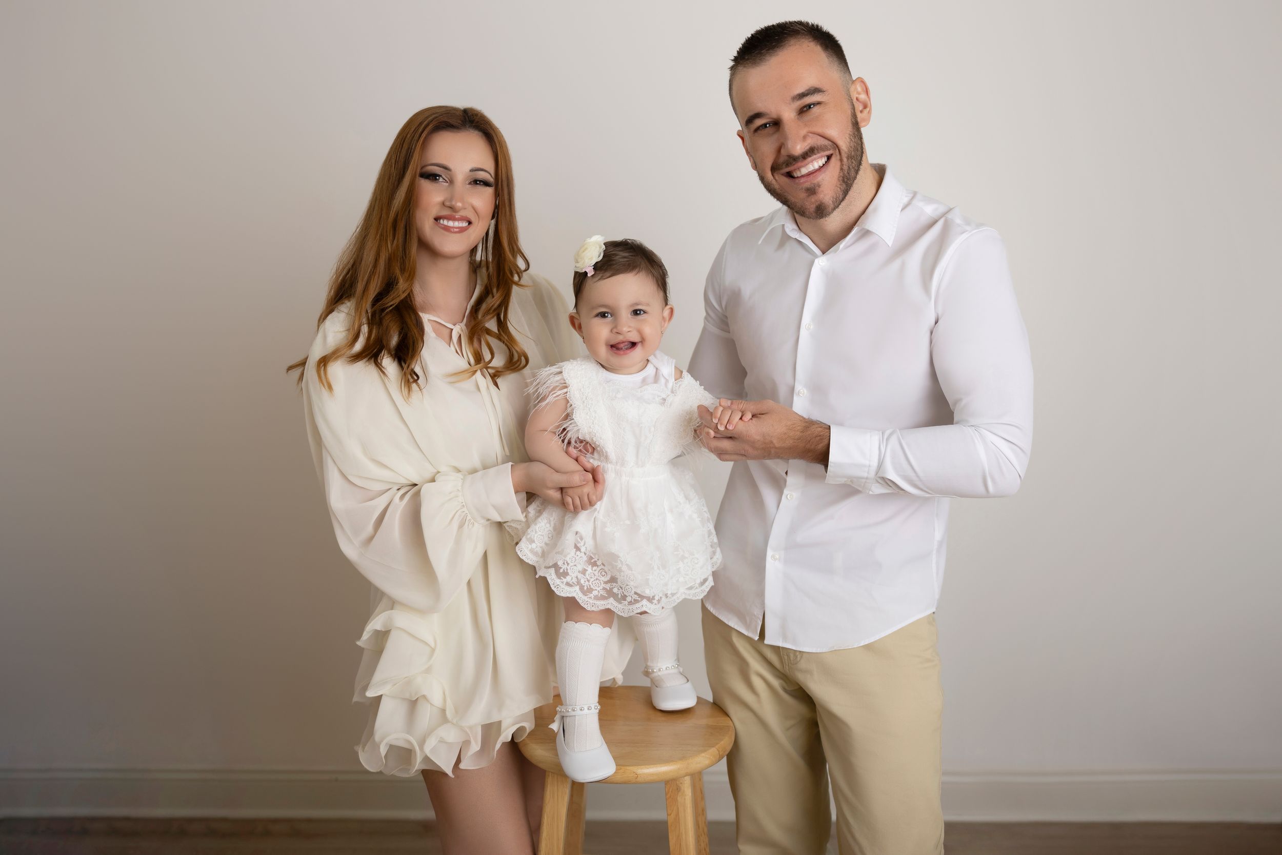 Family in coordinated cream and white outfits poses together against neutral wall for portrait session.