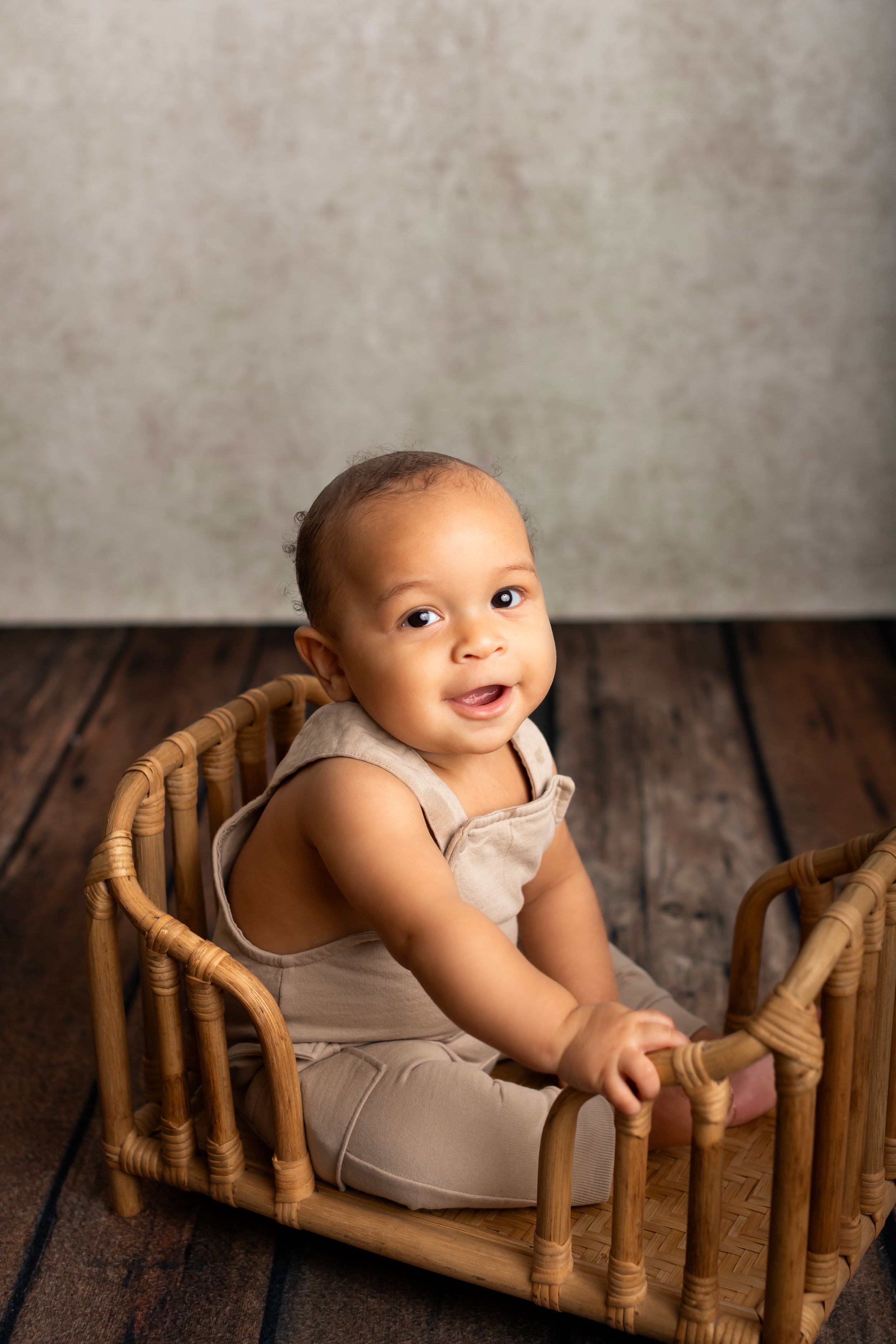 Adorable baby in beige outfit sits smiling in vintage rattan doll bed against grey backdrop and wood floor.
